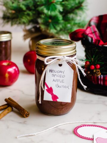A jar of apple butter wrapped with twine and a gift tag with other jars and a Christmas tree in the background.