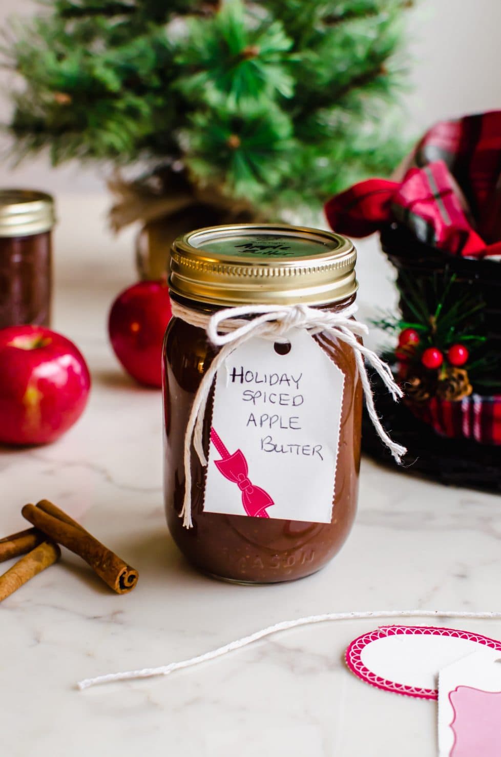 A jar of apple butter wrapped with twine and a gift tag with other jars and a Christmas tree in the background. 