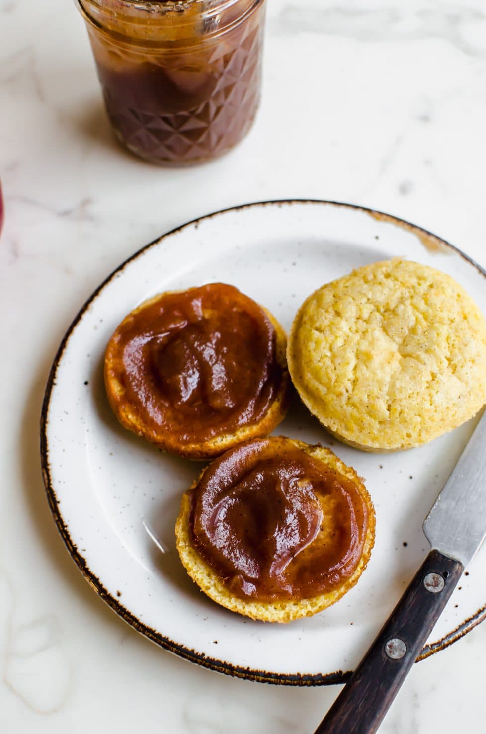 An overhead shot of a white plate with a cornbread muffin sliced open and smeared with apple butter.