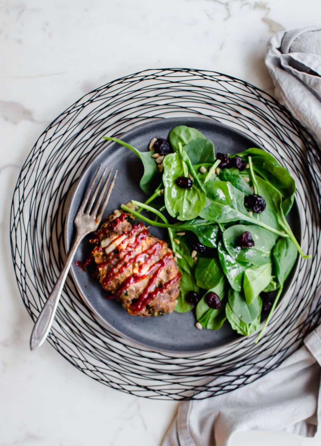 A grey plate on a black wire charger with a serving of mini meatloaf and spinach salad on the side.