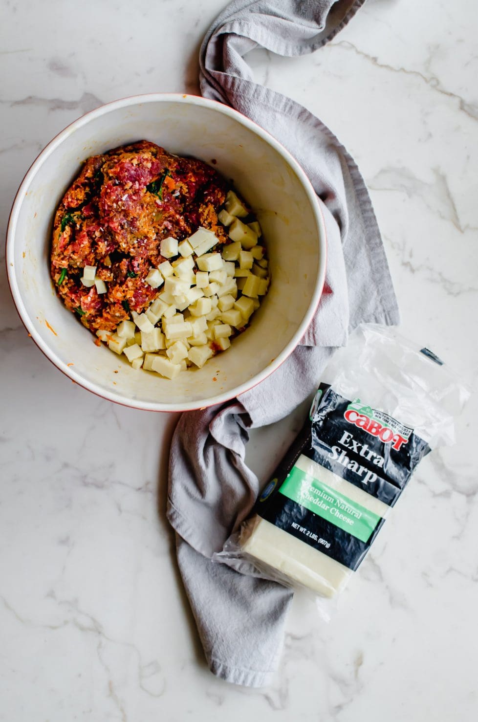 A bowl with meatloaf mixture and cheese cubes inside and a block of white cheddar cheese on the side.