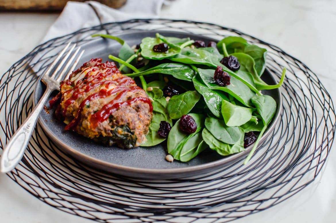 A grey plate on a black wire charger with a serving of mini meatloaf and spinach salad on the side.