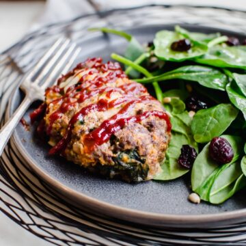 A grey plate on a black wire charger with a serving of mini meatloaf and spinach salad on the side.