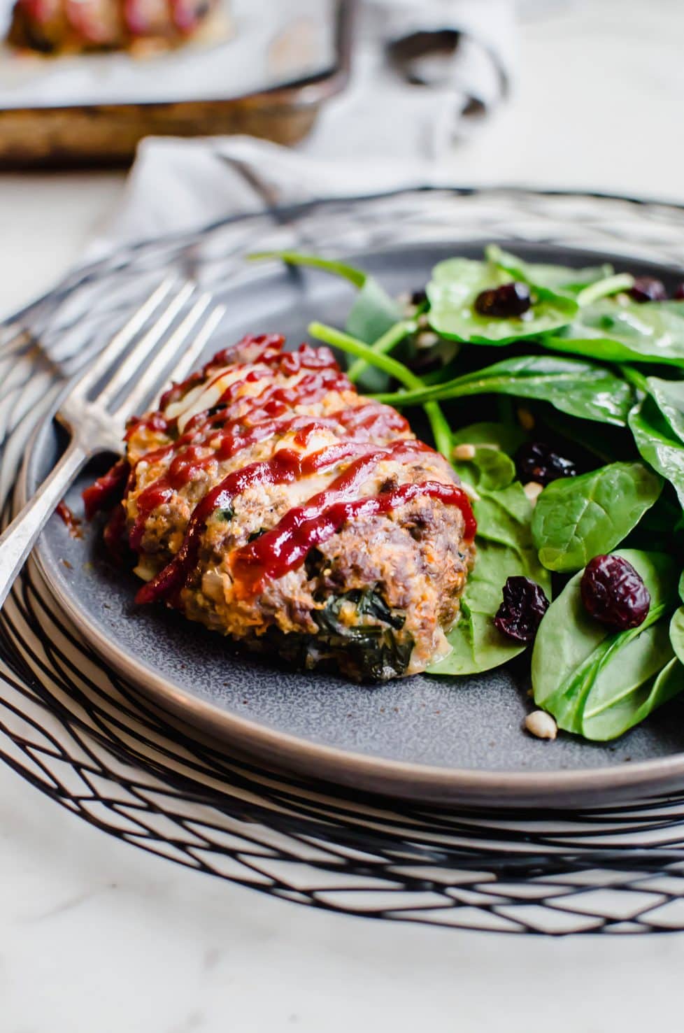 A grey plate on a black wire charger with a serving of mini meatloaf and spinach salad on the side.