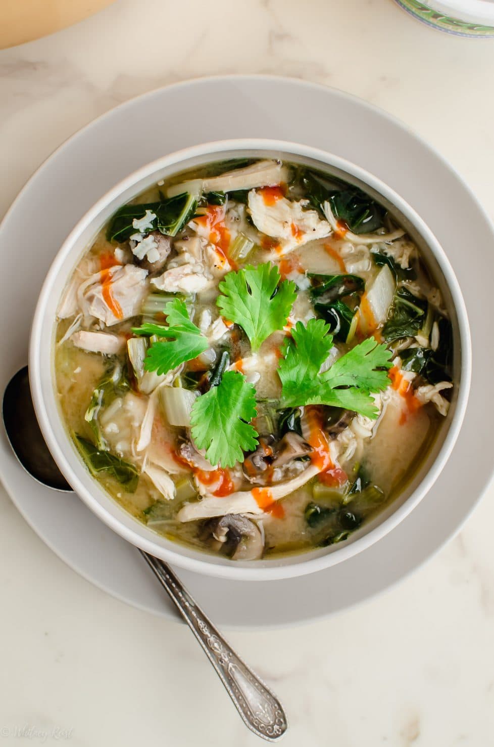 A white bowl filled with miso chicken and rice soup that is garnished with sriracha sauce and a cilantro sprig. 