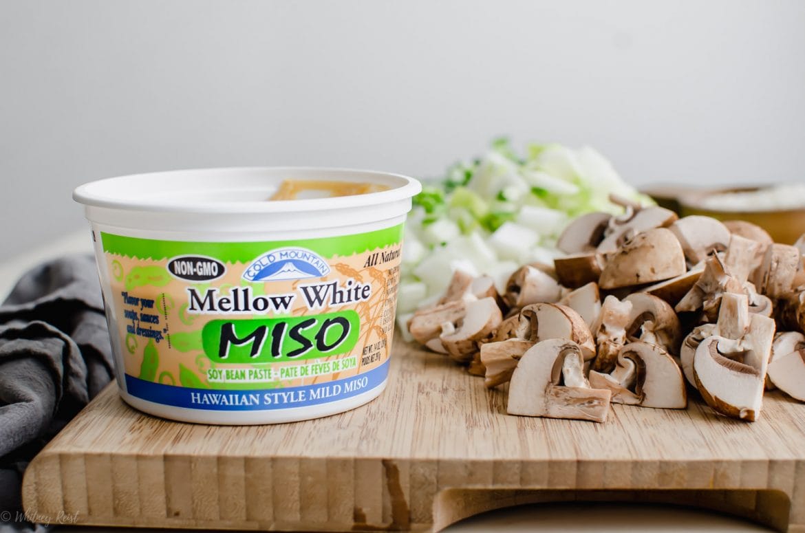 A cutting board topped with chopped mushrooms, bok choy, and a container of miso paste. 