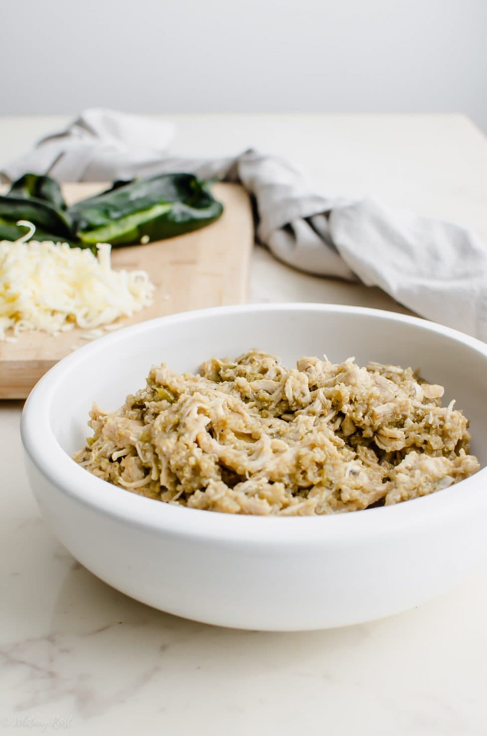 A white bowl filled with quinoa and shredded chicken with a cutting board in the background.