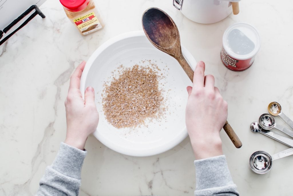 A white bowl with wheat bran inside and two hands holding a wooden spoon next to the bowl. 