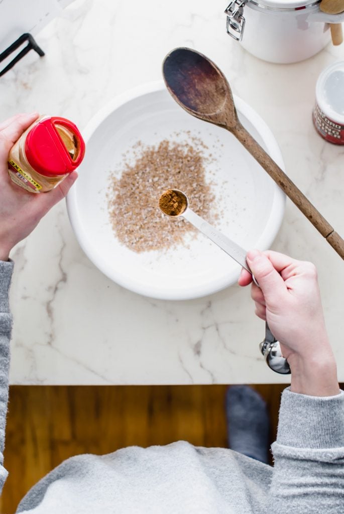 A white bowl with wheat bran in it and hands measuring cinnamon into the bowl.