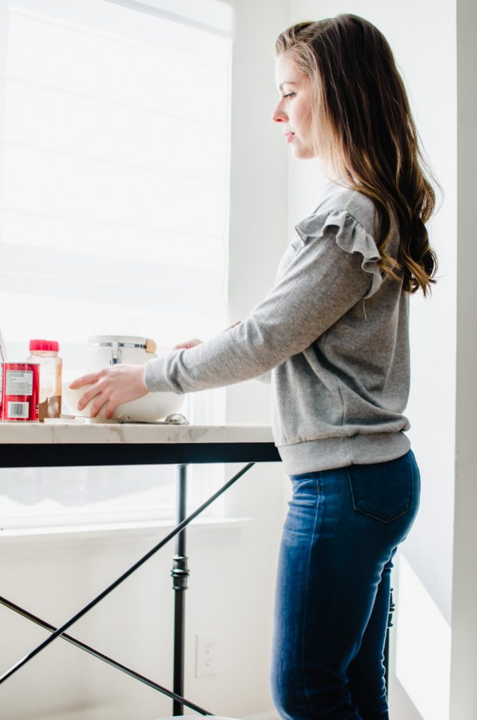 A brunette woman standing a white counter top preparing to mix in a bowl.