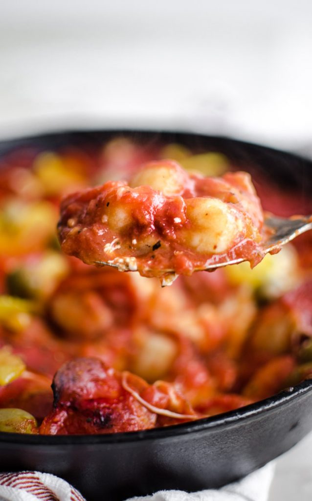 A fork holding up a bite of pizza baked gnocchi with a skillet of the dish in the background.