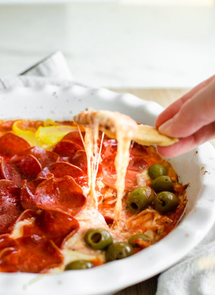 A hand with a pita chip dipping into a dish of baked pizza dip.