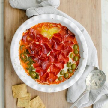 A white round baking dish filled with pizza dip sitting on a cutting board with pita chips.