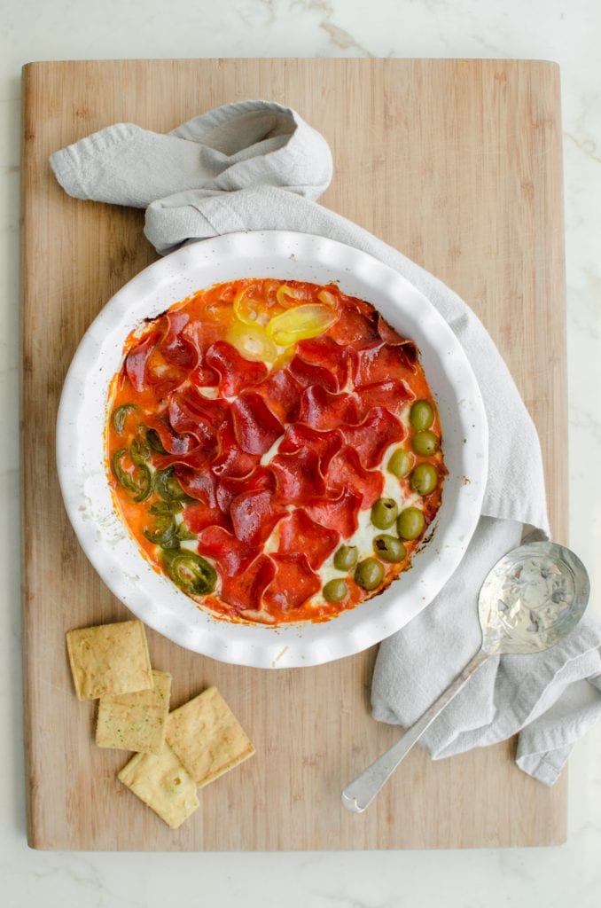 A white round baking dish filled with pizza dip sitting on a cutting board with pita chips.