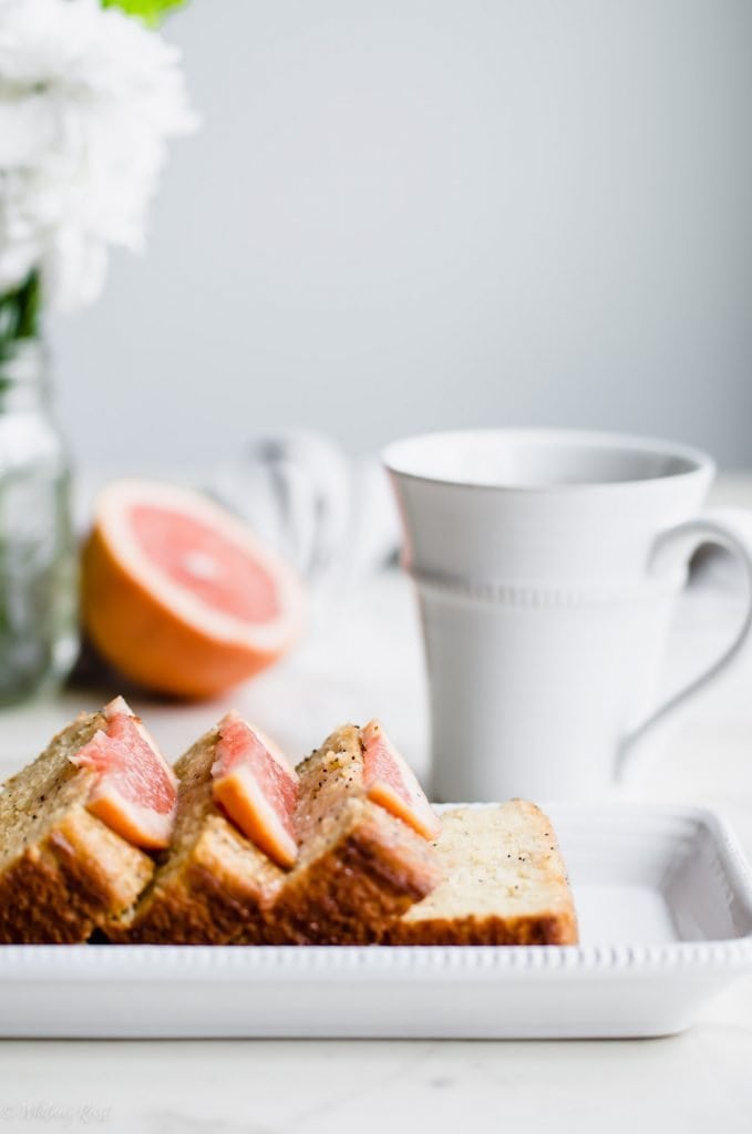 A loaf of sliced grapefruit cake on a white platter with flowers and a cup of tea on the side.