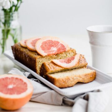 A loaf of sliced grapefruit cake on a white platter with flowers and a cup of tea on the side.