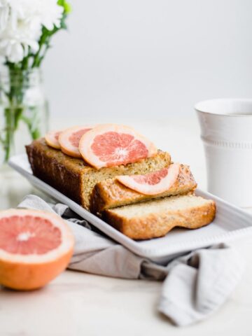 A loaf of sliced grapefruit cake on a white platter with flowers and a cup of tea on the side.