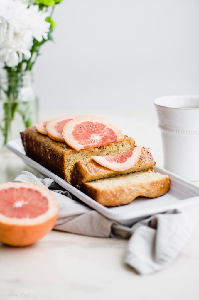 A loaf of sliced grapefruit cake on a white platter with flowers and a cup of tea on the side. 