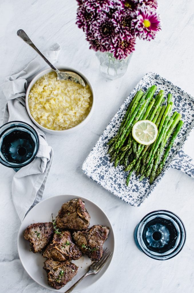 A dinner table set with a spring meal of lamb chops, lemon risotto, and roasted asparagus.