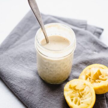 A jar of lemon tahini sauce on a gray towel with juiced lemons on the side.