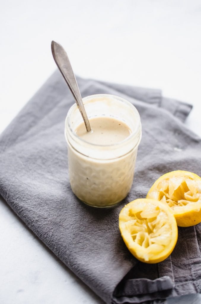 A jar of lemon tahini sauce on a gray towel with juiced lemons on the side. 