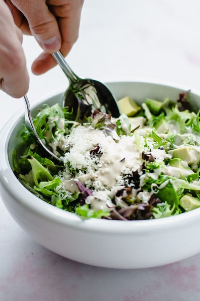 A white bowl filled with springtime everyday salad with hands and utensils tossing the salad.