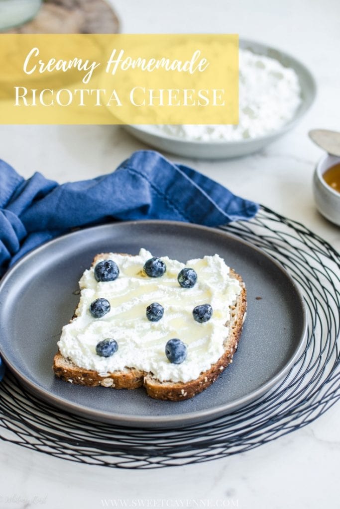 A grey plate with a slice of toast topped with ricotta, honey, and fresh blueberries with a bowl of ricotta in the background.