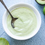 A white bowl filled with creamy avocado ranch dressing on a blue stone counter with limes on the side.
