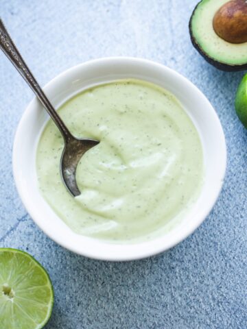 A white bowl filled with creamy avocado ranch dressing on a blue stone counter with limes on the side.