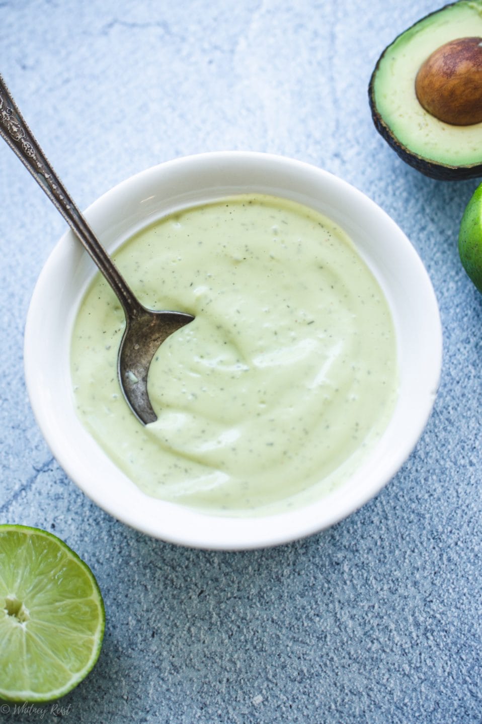 A white bowl filled with creamy avocado ranch dressing on a blue stone counter with limes on the side.