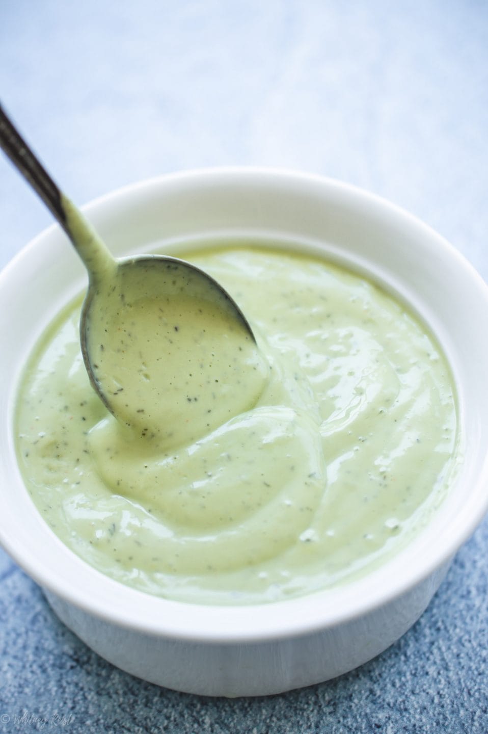 A white bowl filled with creamy avocado ranch dressing on a blue stone counter with limes on the side.