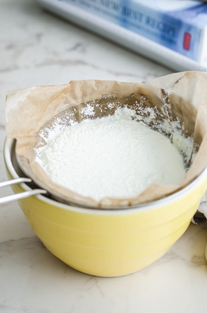 Ricotta being strained through a mesh strainer lined with coffee filters over a yellow bowl on a marble counter top. 