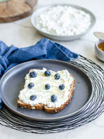 A grey plate with a slice of toast topped with ricotta, honey, and fresh blueberries with a bowl of ricotta in the background.