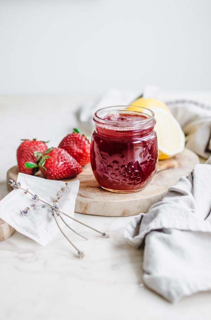 A jar of strawberry lavender syrup on a cutting board with lemons, strawberries, and tea bags on the side.