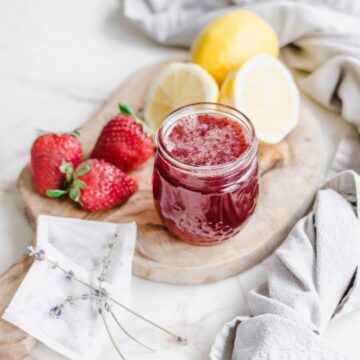 A jar of strawberry lavender syrup on a cutting board with lemons, strawberries, and tea bags on the side.