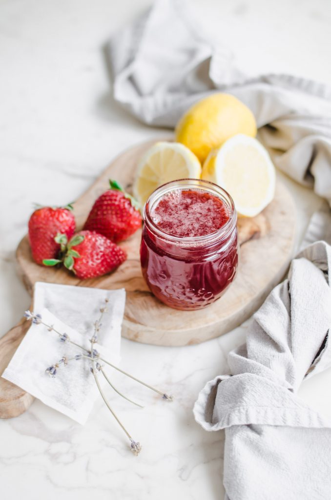 A jar of strawberry lavender syrup on a cutting board with lemons, strawberries, and tea bags on the side.