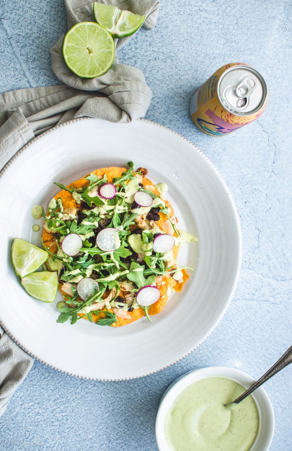 An overhead shot of a chicken tostada on white plate with lime wedges and a bowl of avocado ranch on the side. 