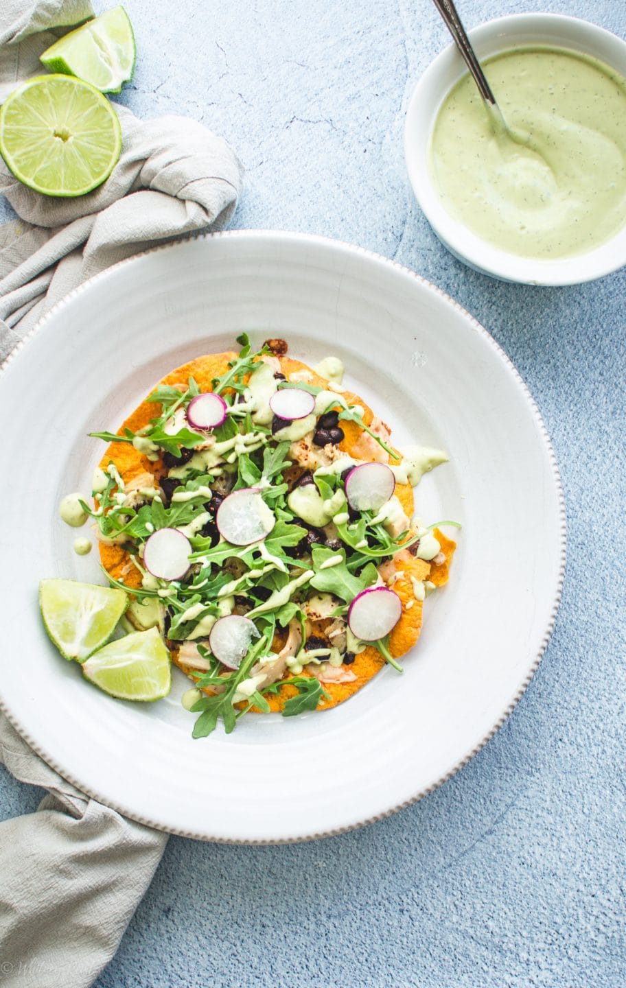 An overhead shot of a chicken tostada on white plate with lime wedges and a bowl of avocado ranch on the side.