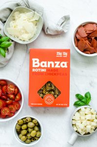 A box of Banza pasta on a white background surrounded by white bowls of ingredients for Pizza Pasta Salad.