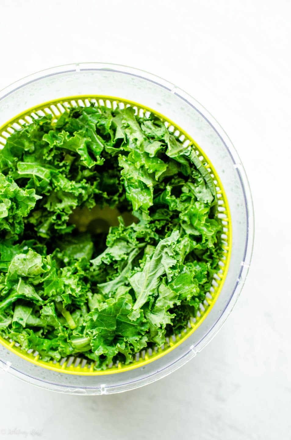 A salad spinner filled with kale on a white stone surface.