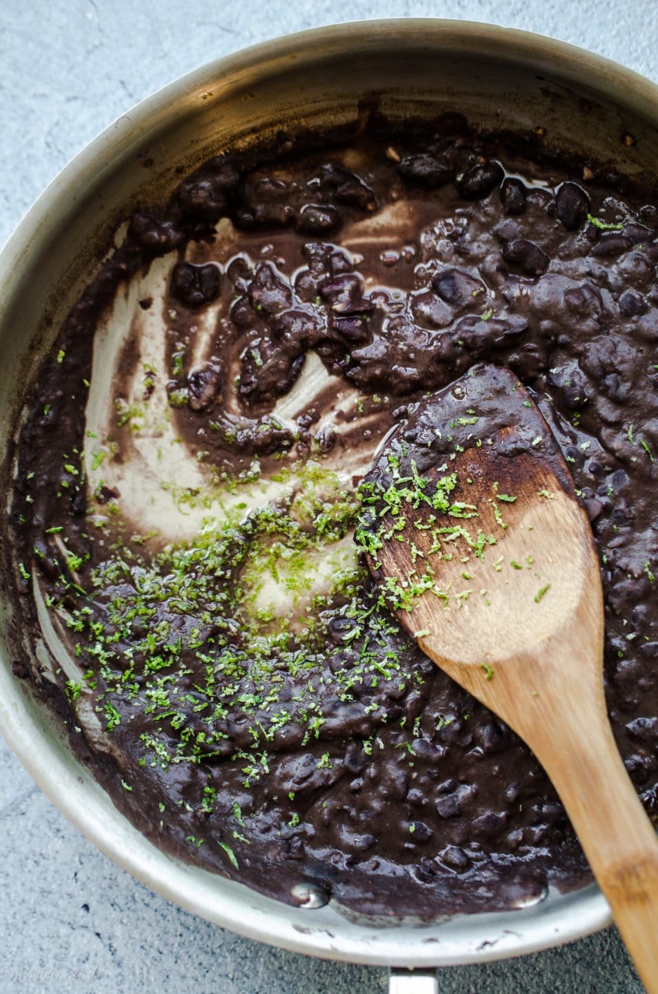 A skillet with mashed black beans being stirred with lime zest with a wooden spoon.