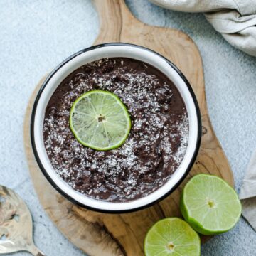 An overhead shot of refried black beans in a white bowl topped with cotija cheese and a lime slice.