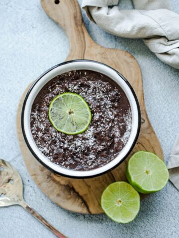 An overhead shot of refried black beans in a white bowl topped with cotija cheese and a lime slice.