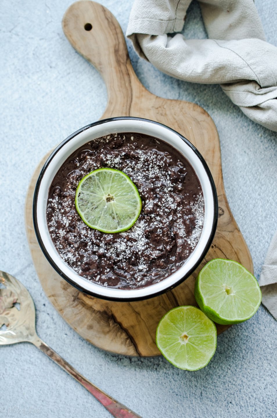 An overhead shot of refried black beans in a white bowl topped with cotija cheese and a lime slice.