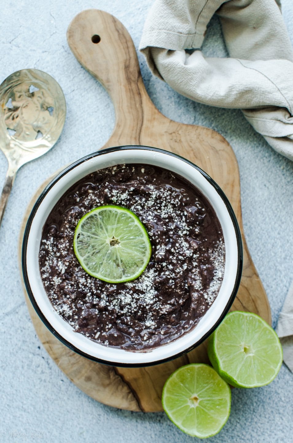 An overhead shot of refried black beans in a white bowl topped with cotija cheese and a lime slice.