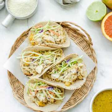 An overhead shot of a wicker charger with parchment paper and Mango Mojo Chicken Tacos with a side of mojo sauce and avocado ranch on the side.