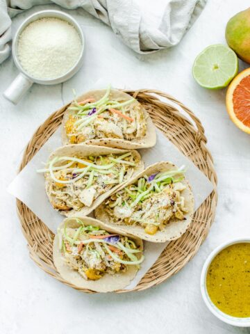 An overhead shot of a wicker charger with parchment paper and Mango Mojo Chicken Tacos with a side of mojo sauce and avocado ranch on the side.
