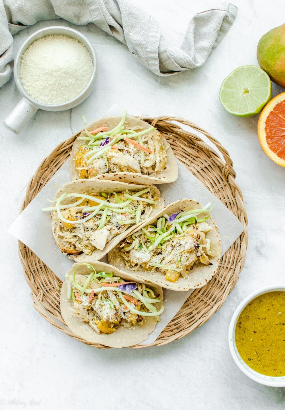 An overhead shot of a wicker charger with parchment paper and Mango Mojo Chicken Tacos with a side of mojo sauce and avocado ranch on the side.