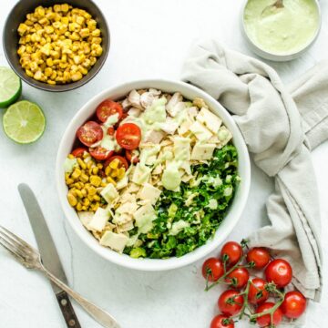 An overhead shot of a white bowl filled with chopped Mexican kale salad with bowls of the ingredients on the side.