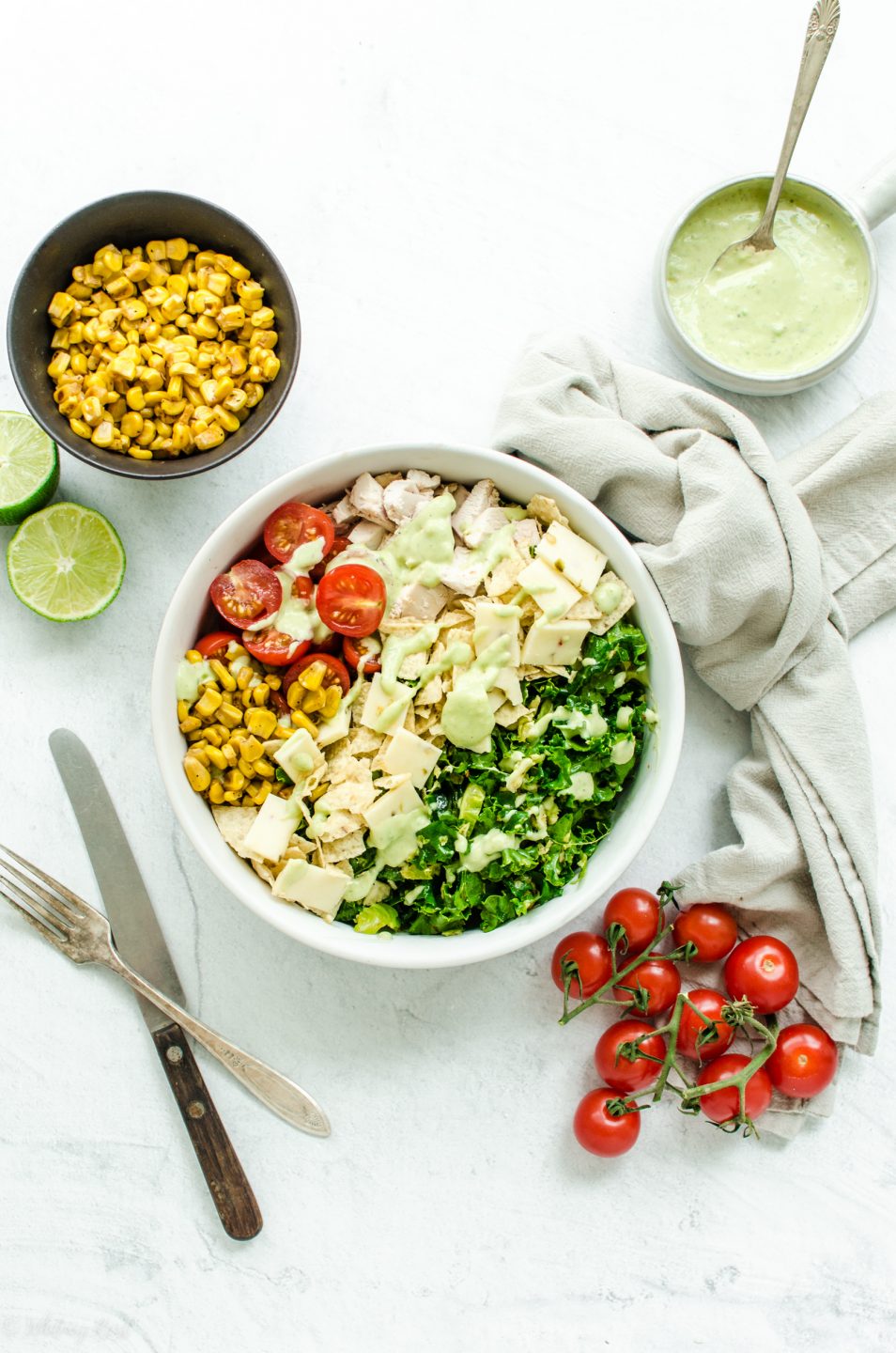 An overhead shot of a white bowl filled with chopped Mexican kale salad with bowls of the ingredients on the side.